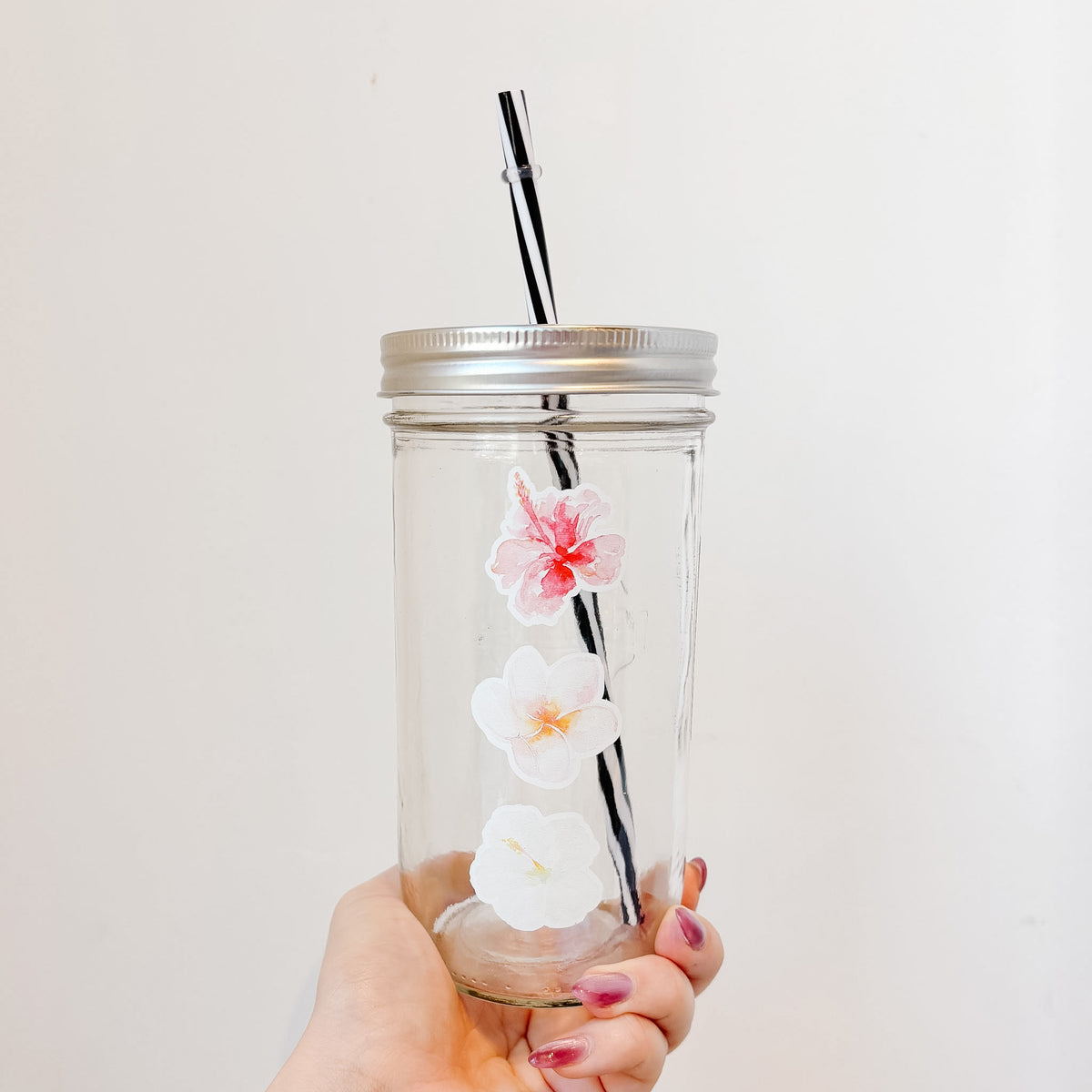 A hand holding a clear glass mason jar tumbler with a silver metal lid and a black-and-white striped straw. The jar is decorated with a pink, peach, and white hibiscus flower. 