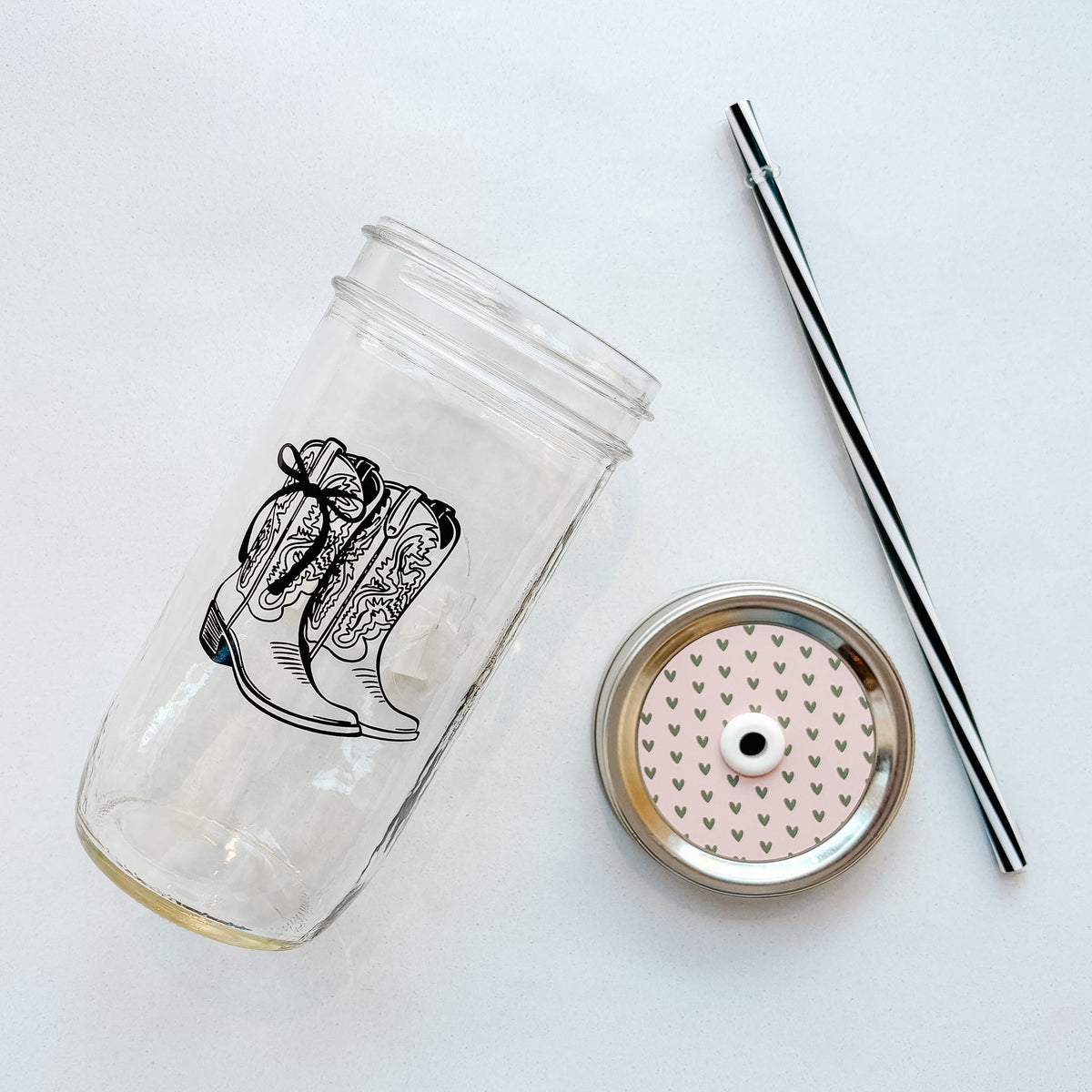 A clear glass tumbler with a black cowboy boot illustration with a bow, shown with a black-and-white striped straw and a metal straw lid feating gold hearts on a pale pink background.