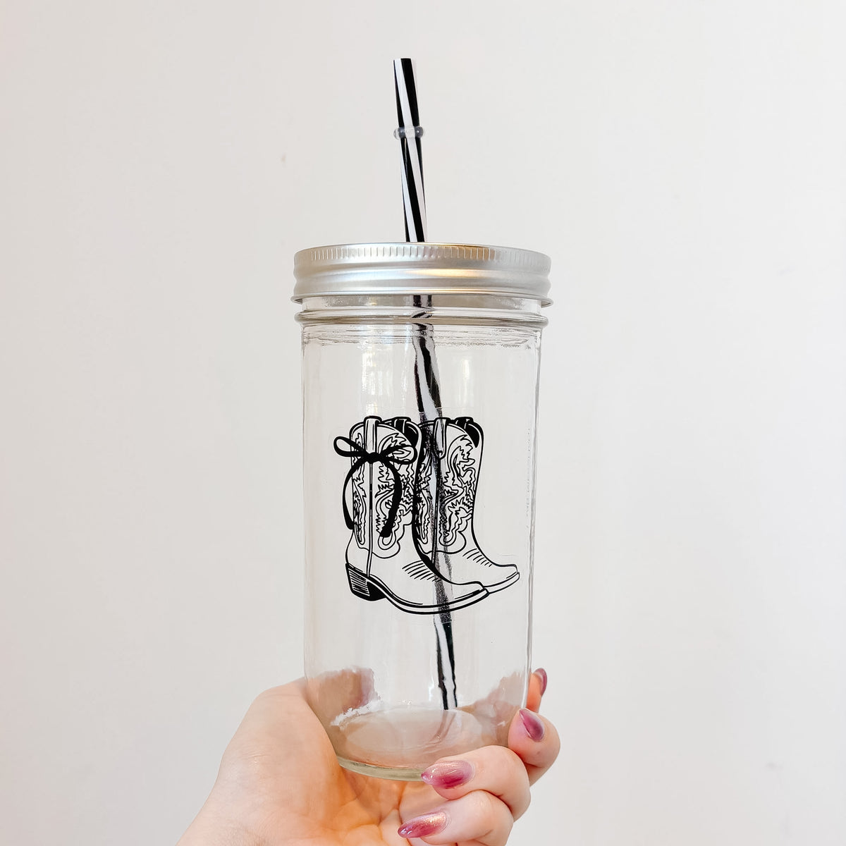 A hand holding a clear glass mason jar tumbler with a silver metal lid and a black-and-white striped straw. The glass features a black line art illustration of a western cowboy boot with a bow.