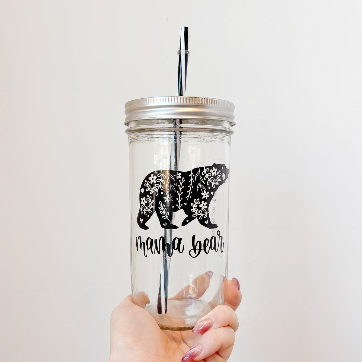 A hand holding a clear glass mason jar tumbler with a silver metal lid and black straw. The jar features a black floral bear silhouette and the text 'mama bear' in black script.