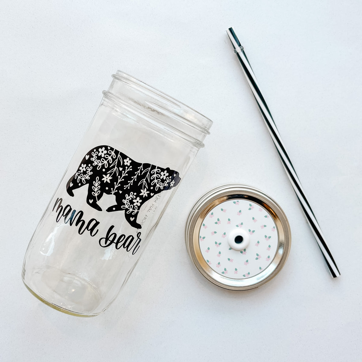 A clear glass tumbler with a black floral bear silhouette and the words 'mama bear' printed on it, shown with a metal lid and a black-and-white striped straw on a white background.