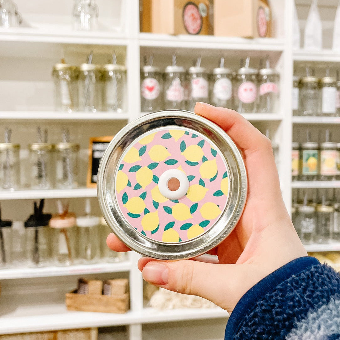 A hand holding a mason jar patterned straw lid with lemons on a light pink background