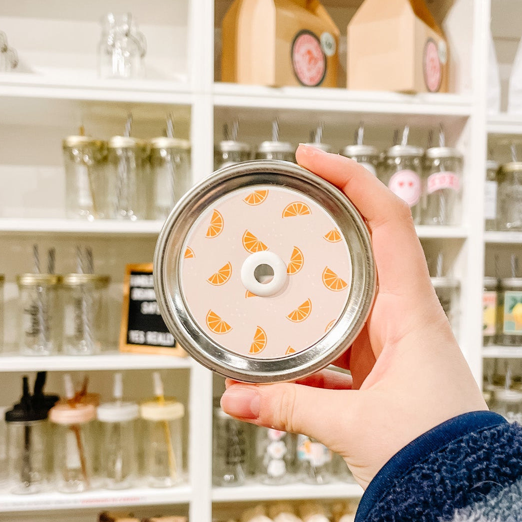 Hand holding a mason jar patterned straw lid with little orange slices on a light peach background