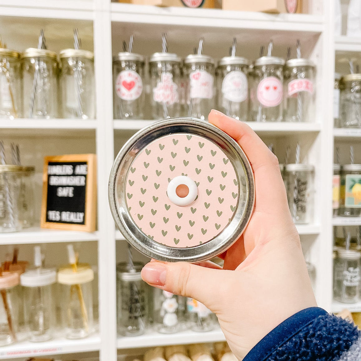 Hand holding mason jar patterned straw lid with small green hearts on a light pink background