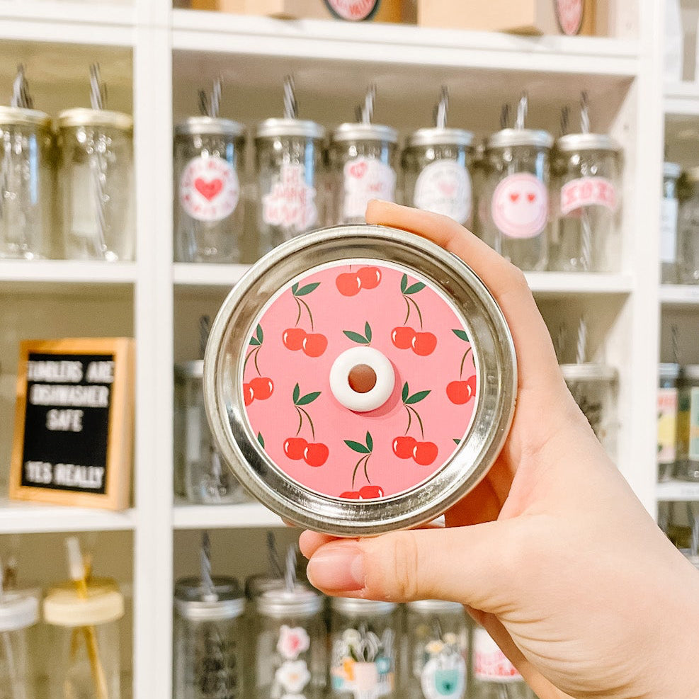 Hand holding a pink lid with cherry design in front of shelves with jars.