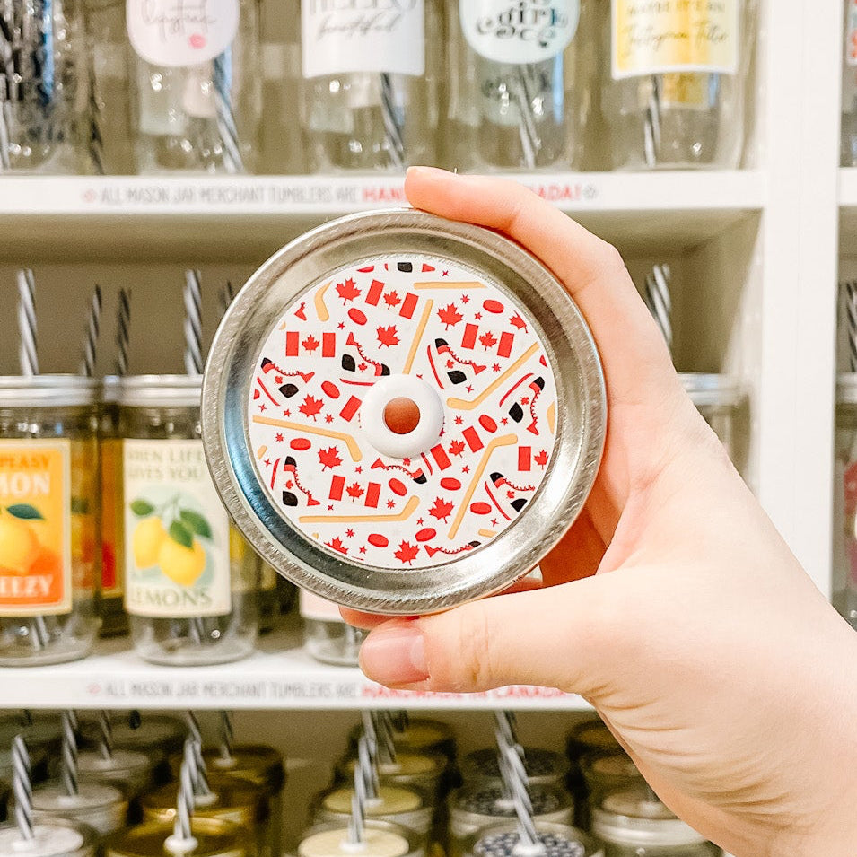 Hand holding mason jar patterned straw lid with hockey designs including skates, sticks, pucks and Canada flag on white background 
