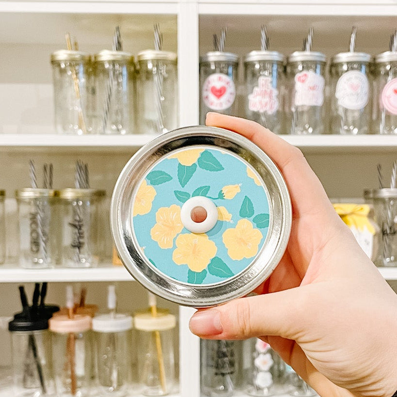 Hand holding a mason jar patterned straw lid That has yellow bright flowers on a blue background 