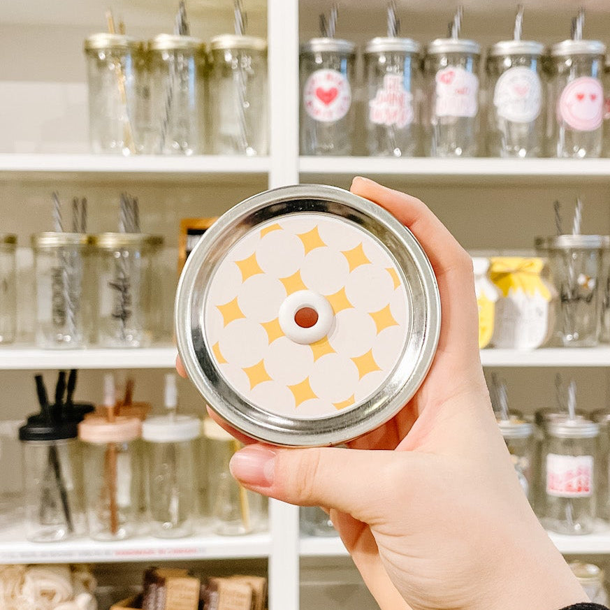 Hand holding a mason jar patterned straw lid That has yellow diamond shapes on an off white background 