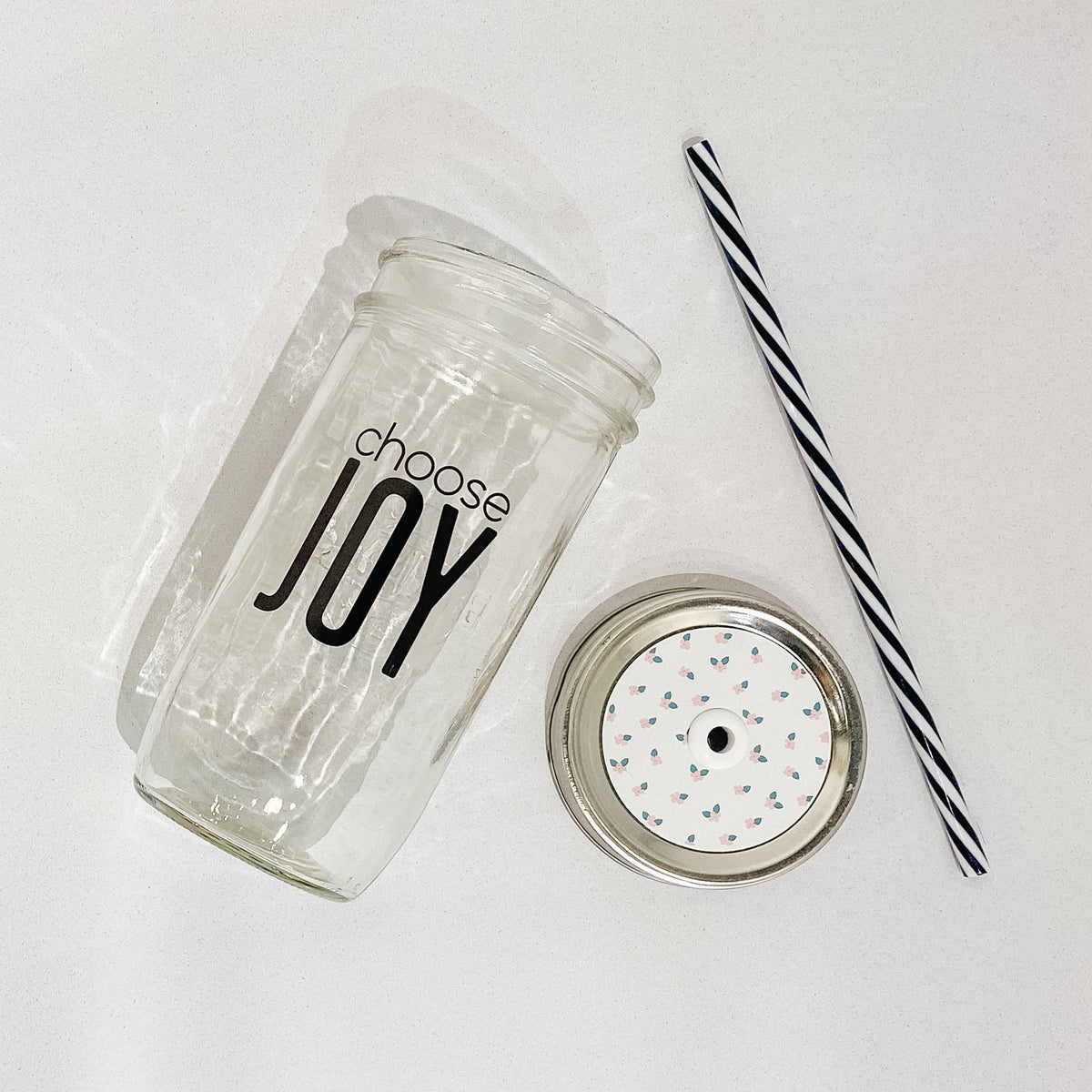 Flat lay of mason jar tumbler that reads choose joy in black ink with clear background. Black and white striped plastic straw and a mason jar patterned straw lid with small pink flowers on white background. 