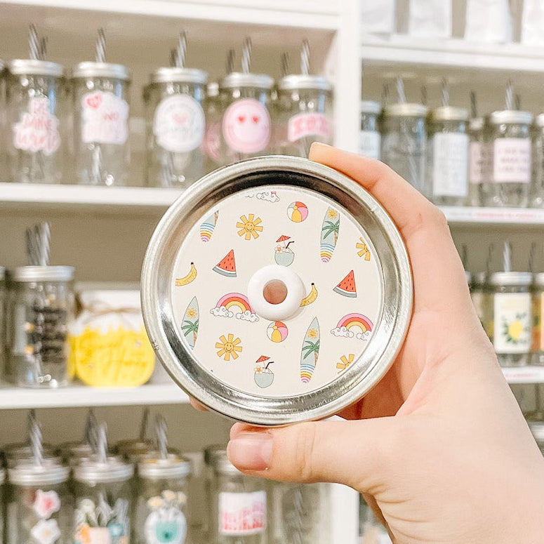 Hand holding mason jar patterned straw lid that has assorted watermelon, surf boards, rainbows, beach balls, drinks and suns on beige off white background 