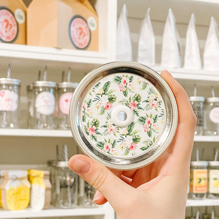 Hand holding mason jar patterned straw lid that has mixed floral arrangements that are tropical on white background 