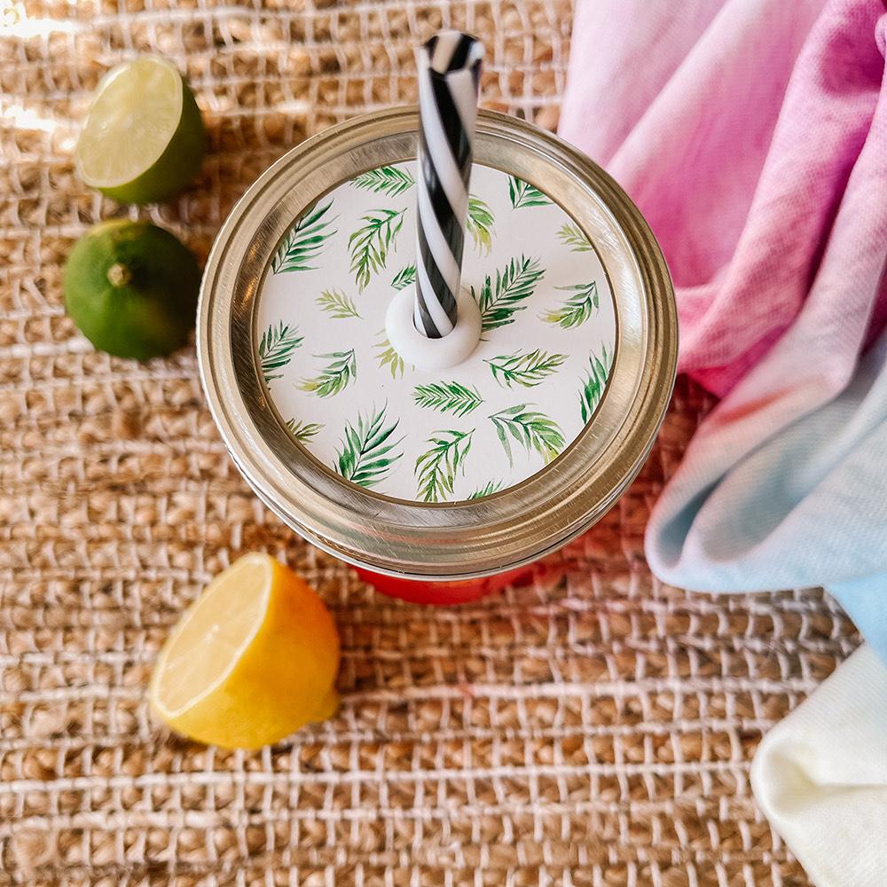 Mason jar straw lid with silver ring and a white sticker that has palm leaves in print. Photographed as a flat lay in a weave mat with a scarf and some lemon slices.