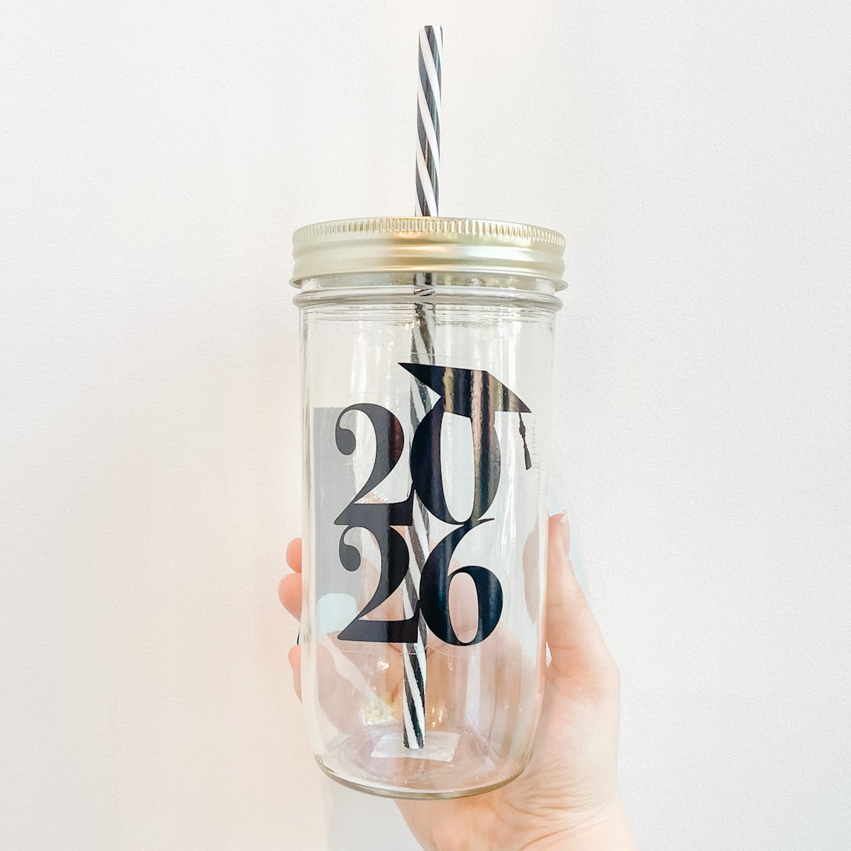Hand holding a mason jar tumbler with a black design of bold 2026 numbers with a small grad cap hanging off the 0. Black and white reusable straw and straw lid 