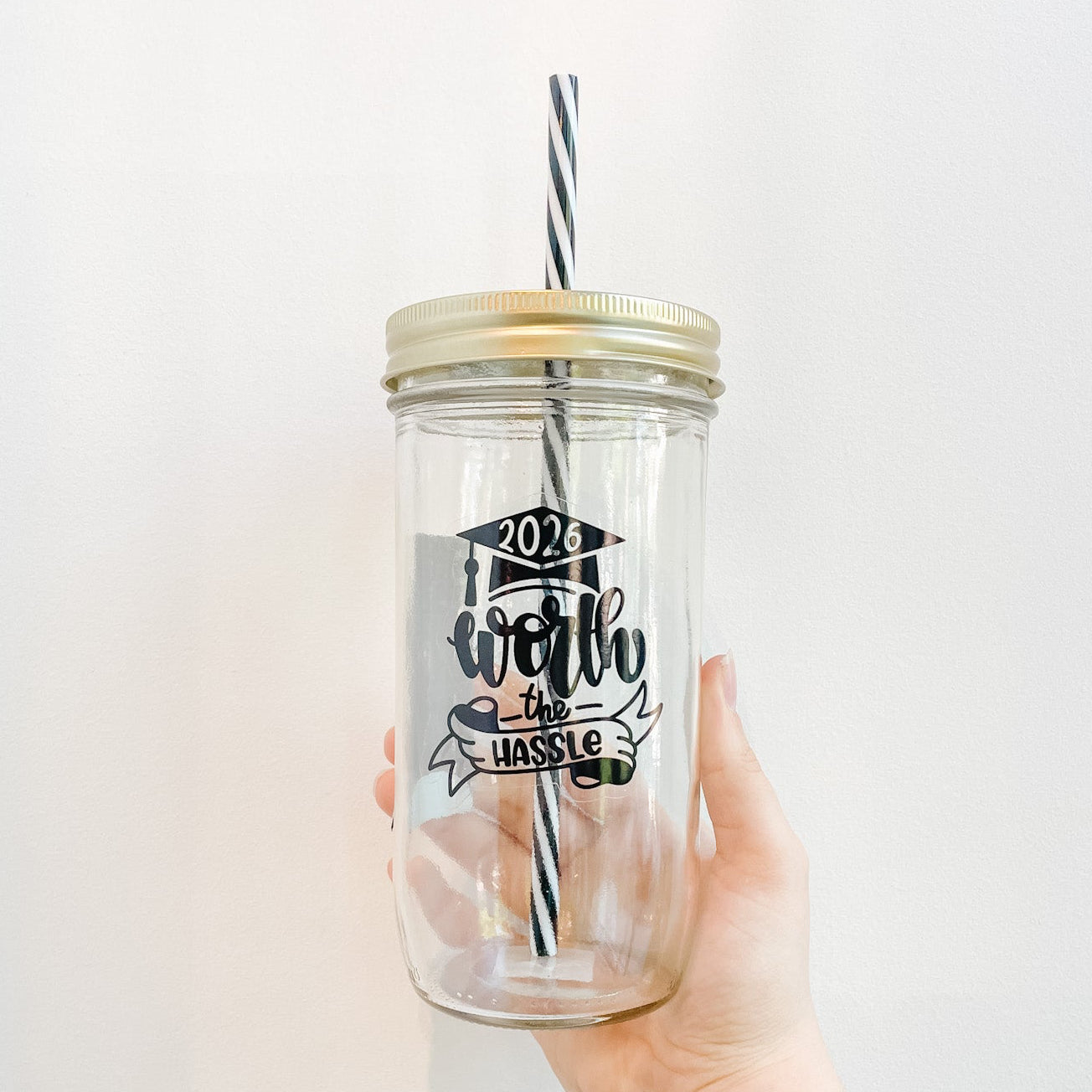 Hand holding a mason jar tumbler that reads in black text 2026 worth the hassle including a banner and grad cap design. Black and white reusable striped straw and straw lid. 
