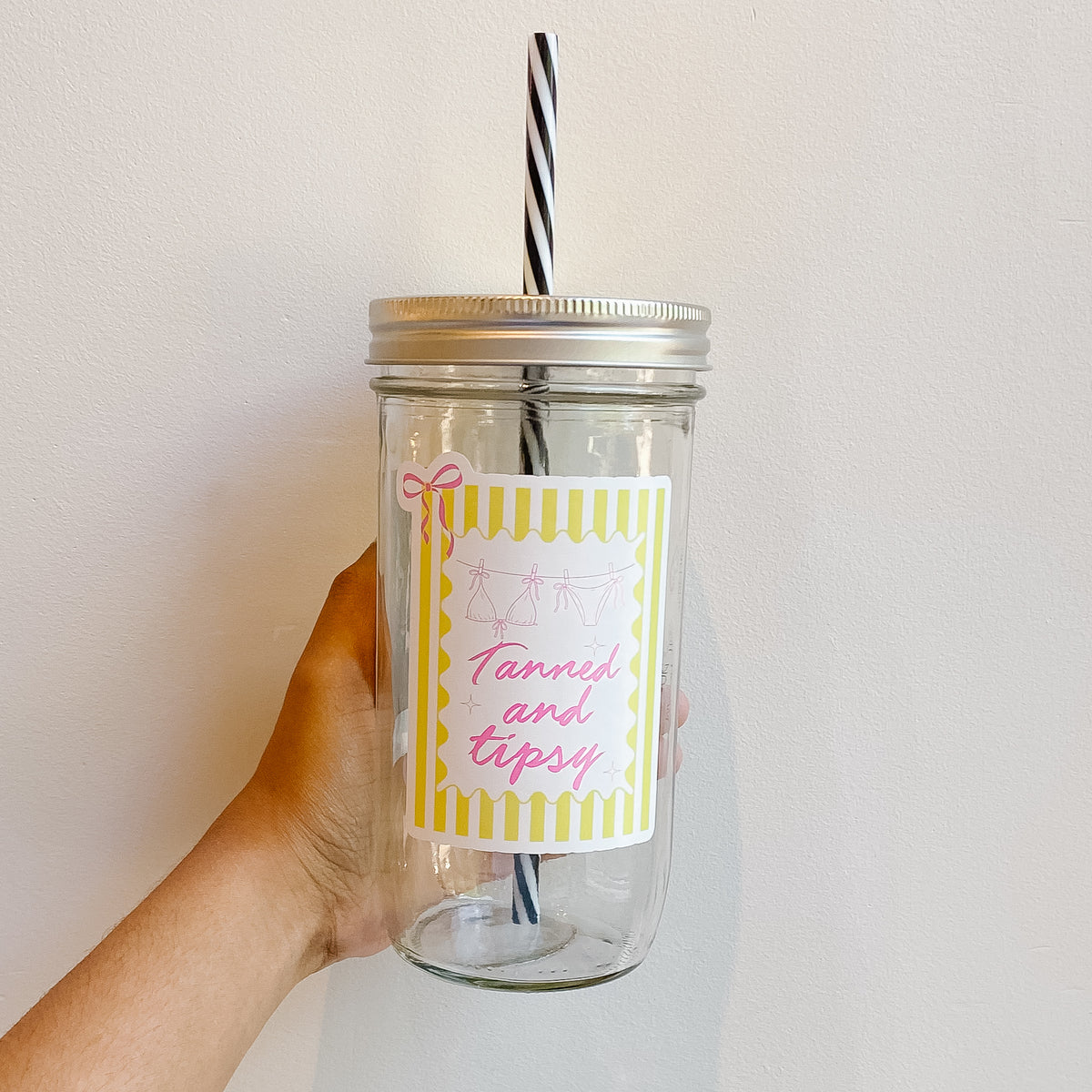 Hand holding mason jar tumbler with yellow striped design that reads tanned and tipsy with pink bikini and bow designs 
