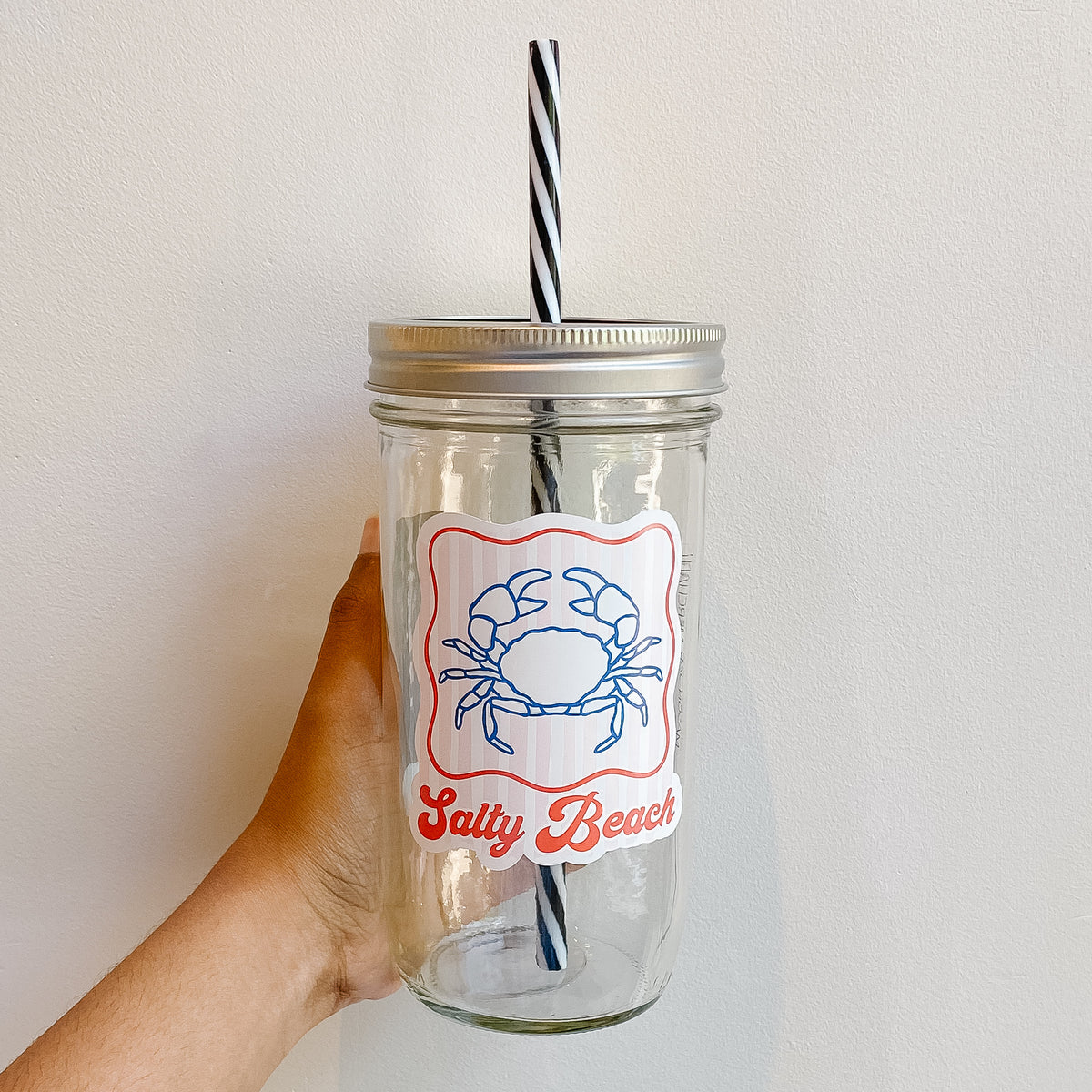 Hand holding a mason jar tumbler that reads salty beach in red writing with a crab and light pale pink/white striped background and red border 