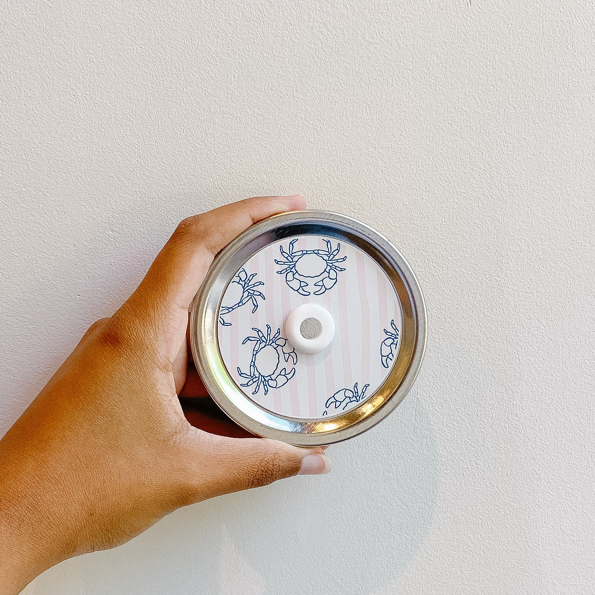 Hand holding a mason jar patterned straw lid that has a light pale pink and white striped background with blue outlined crab designs