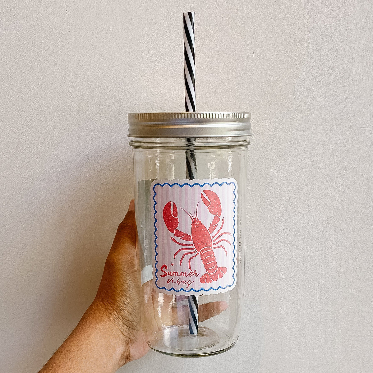 Hand holding a mason jar tumbler with lobster design that reads summer vibes and pink/white striped background with blue border 