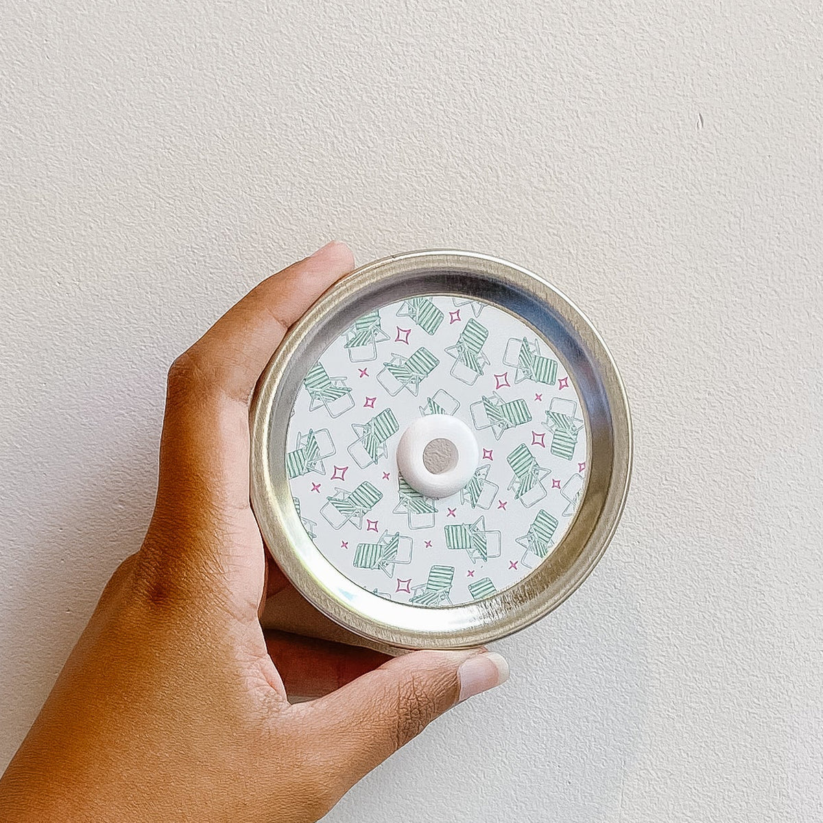 Hand holding a mason jar patterned straw lid that has small green lawn chairs over lid and red outlined star designs on white background 