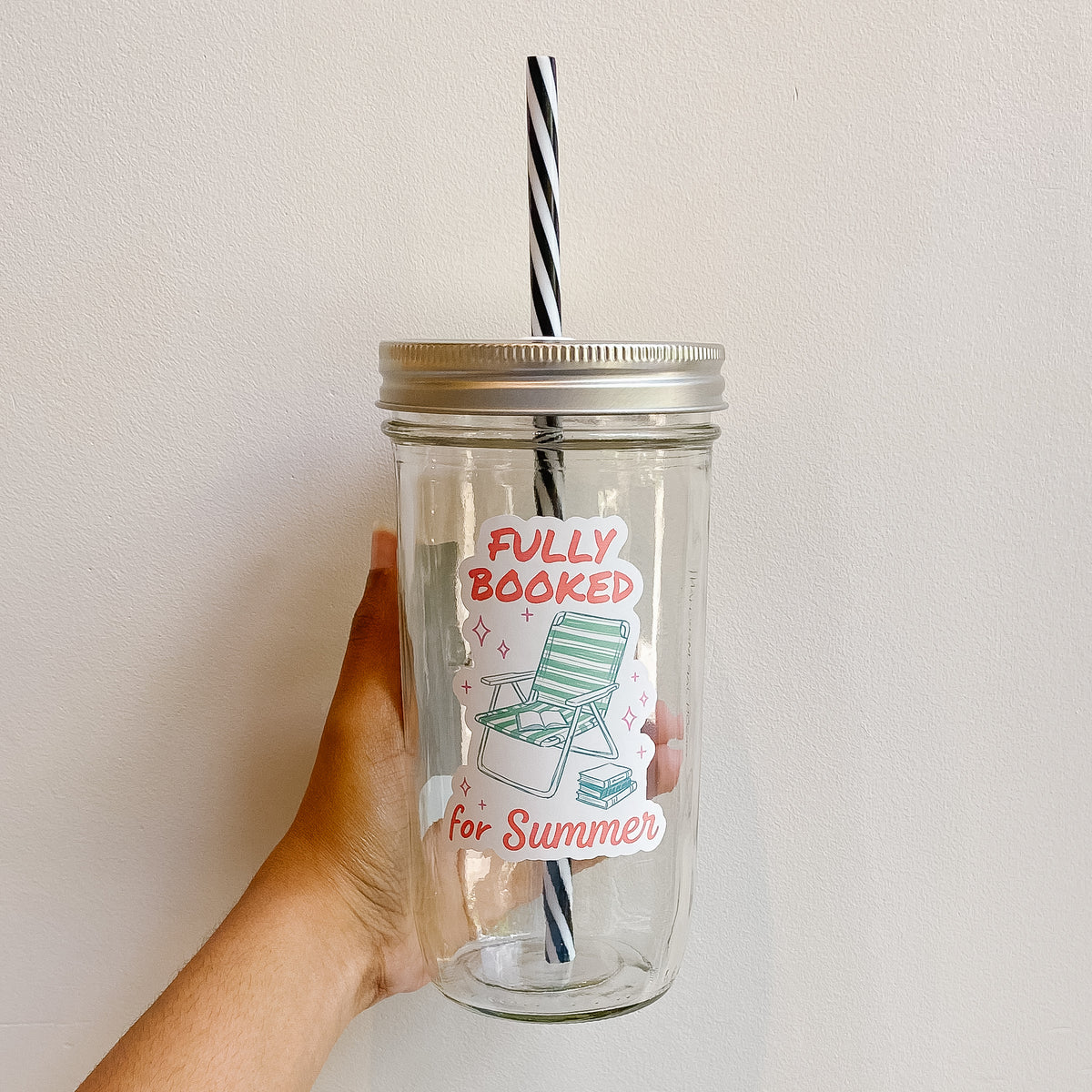 Hand holding a mason jar tumbler that reads fully booked for summer in red ink with a green lawn chair and books stacked  on whiter background 