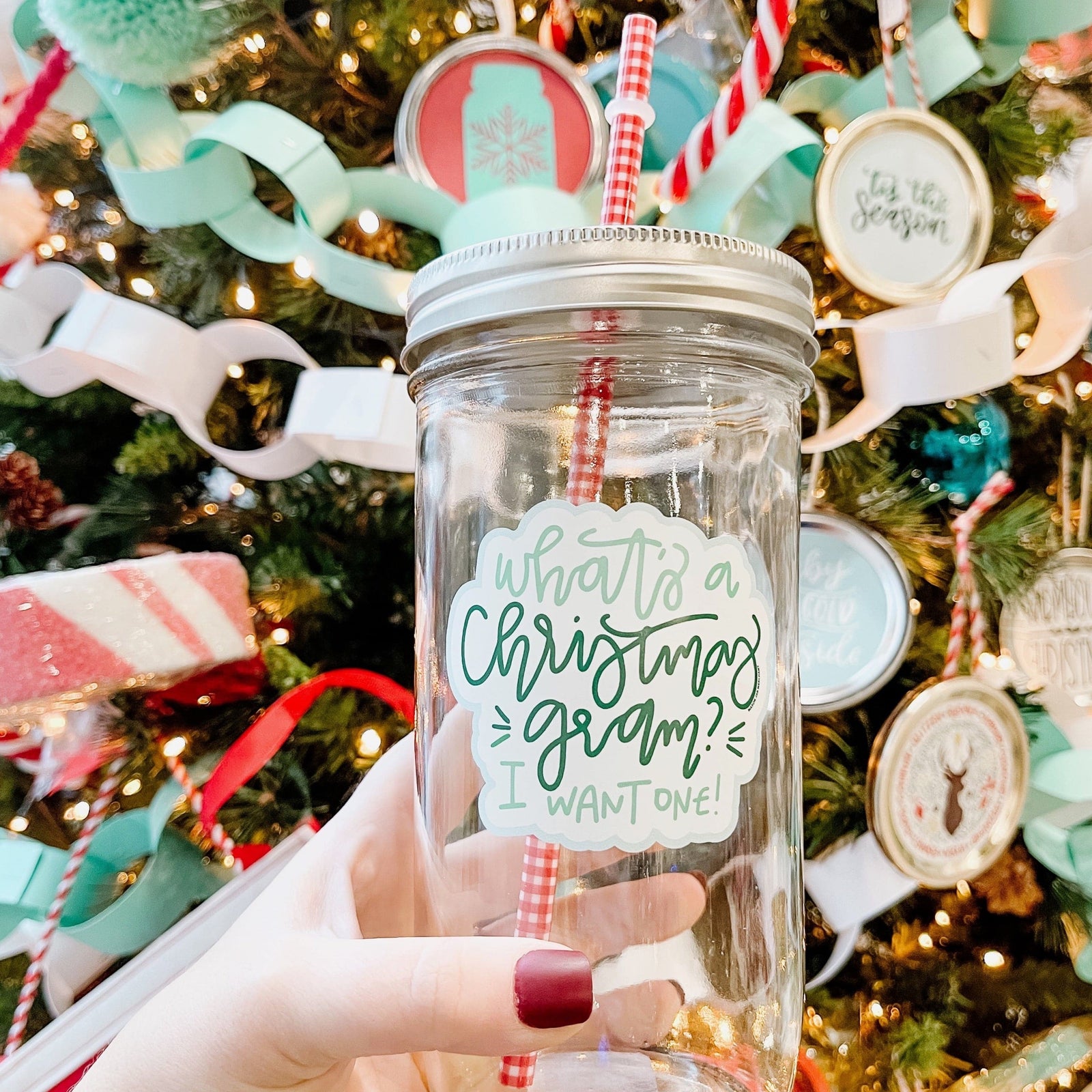 A woman holding an Elf Theme regular mouth mason jar tumbler with silver lid and red and white pattern straw. The tumbler has a print that says "What's a Christmas gram? I want one" in a green color. Behind it is a Christmas tree.