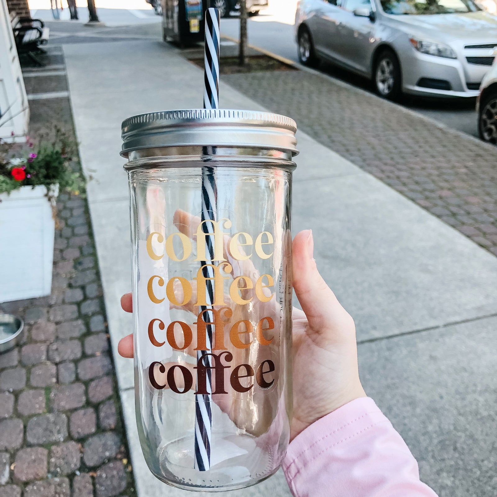 Flat lay of a mason jar tumbler that says “coffee” 4 times in different shades of brown, a straw lid in gold, and a black and white striped straw.