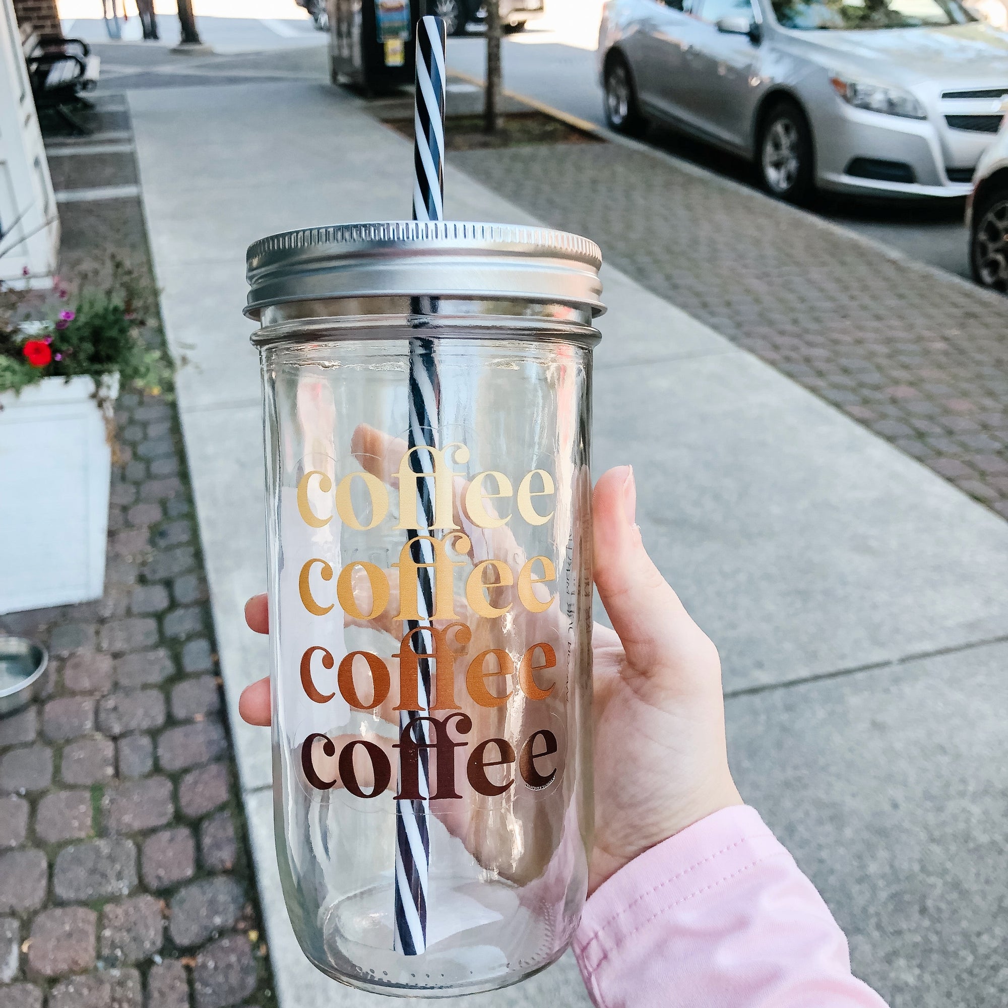 Flat lay of a mason jar tumbler that says “coffee” 4 times in different shades of brown, a straw lid in gold, and a black and white striped straw.