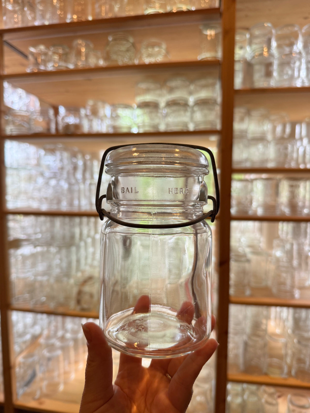 Hand holding a vintage 1930s unmarked Hazel Atlas wire bail pint mason jar with clear glass and dimple neck, store shelves of jars behind