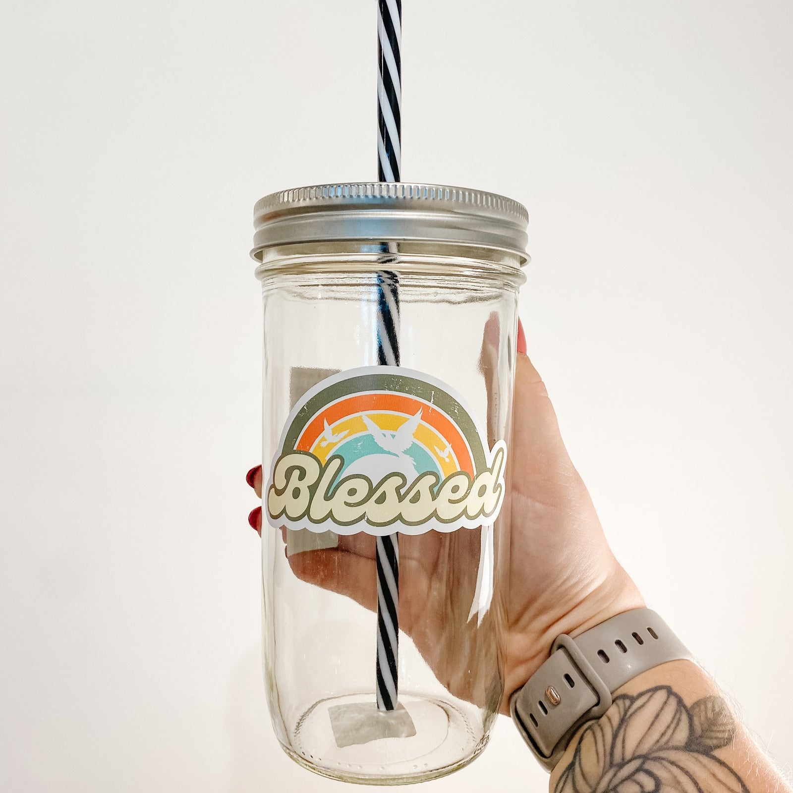 Hand holding a mason jar tumbler that says "Blessed" with a rainbow and white silhouettes of birds.