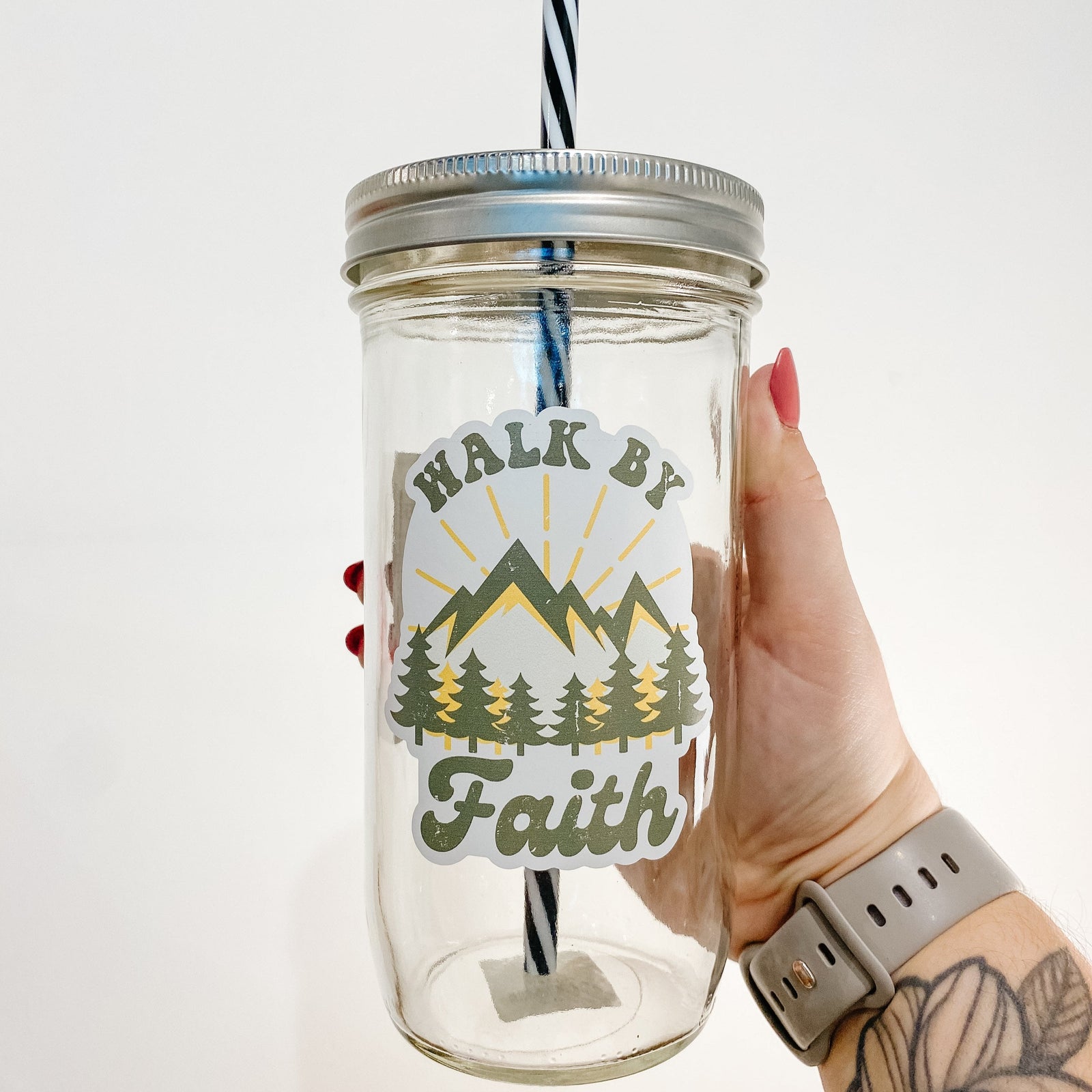 Hand holding a mason jar tumbler that says "walk by faith" with mountains and trees (green and yellow).