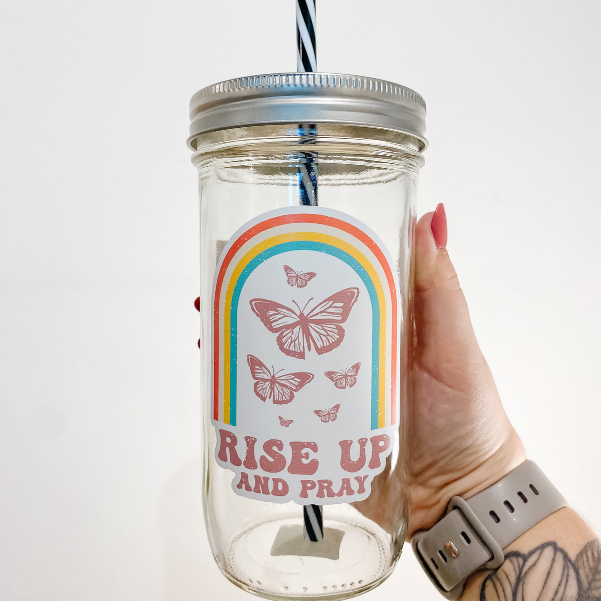 Hand holding mason jar tumbler that says &quot;Rise up and pray” with a rainbow and butterflies