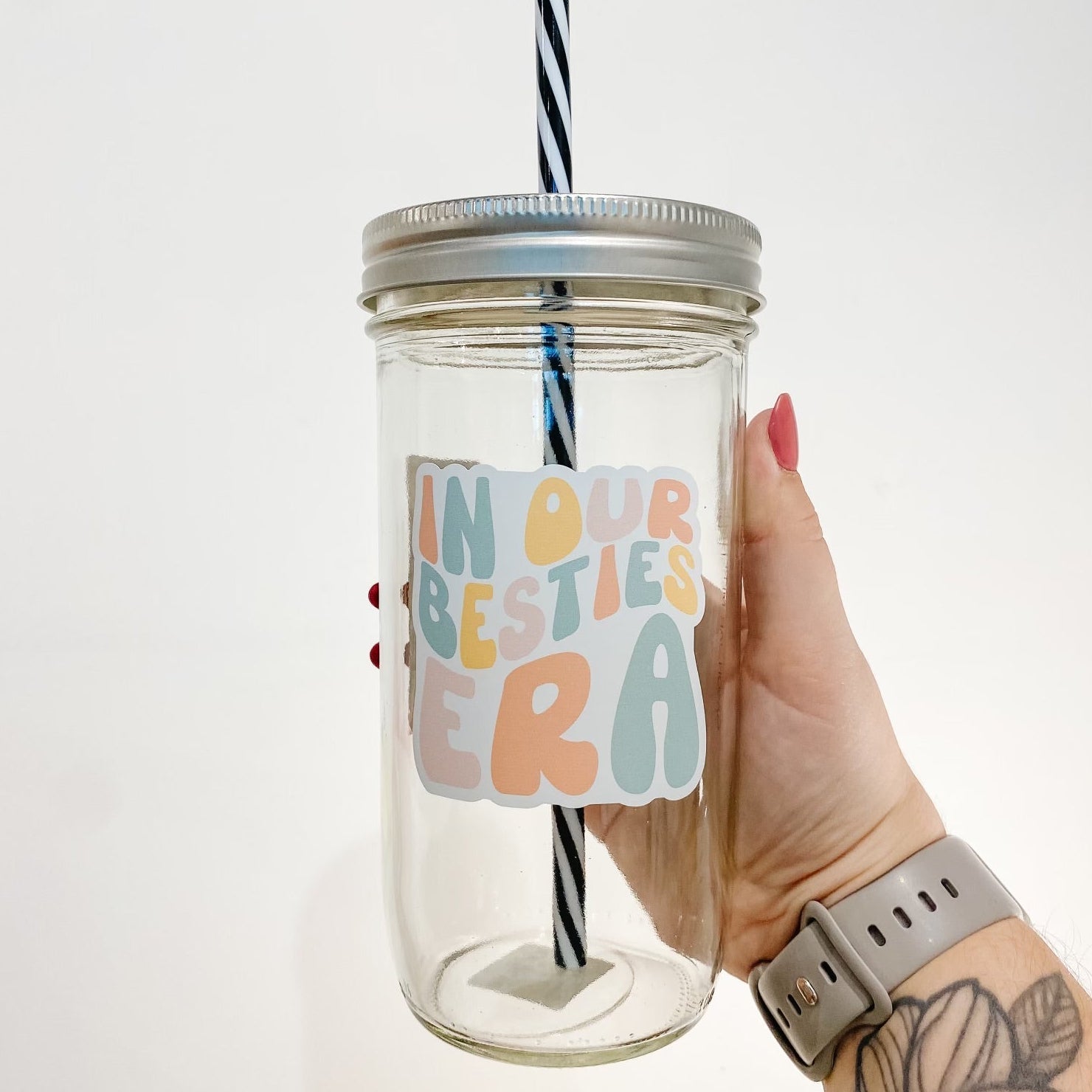 hand holding a mason jar tumbler that reads "in our besties era" the lettering is bubbly and in different shades of yellow, orange, pink, and blue