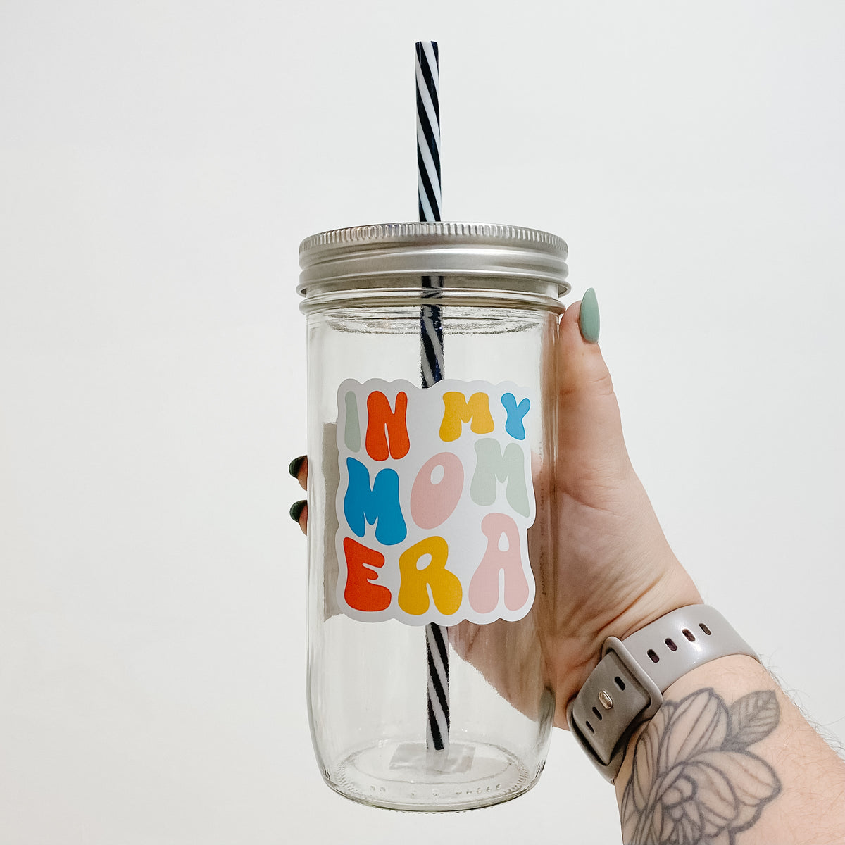 Hand holding a mason jar tumbler that says &quot;In my mom era&quot; in bubbly, different colored letters. 