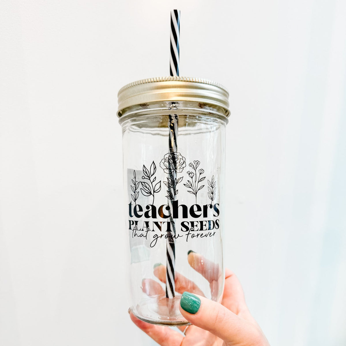 Clear tumbler with gold lid and black and white straw, held by a hand with green nail polish against a light background.