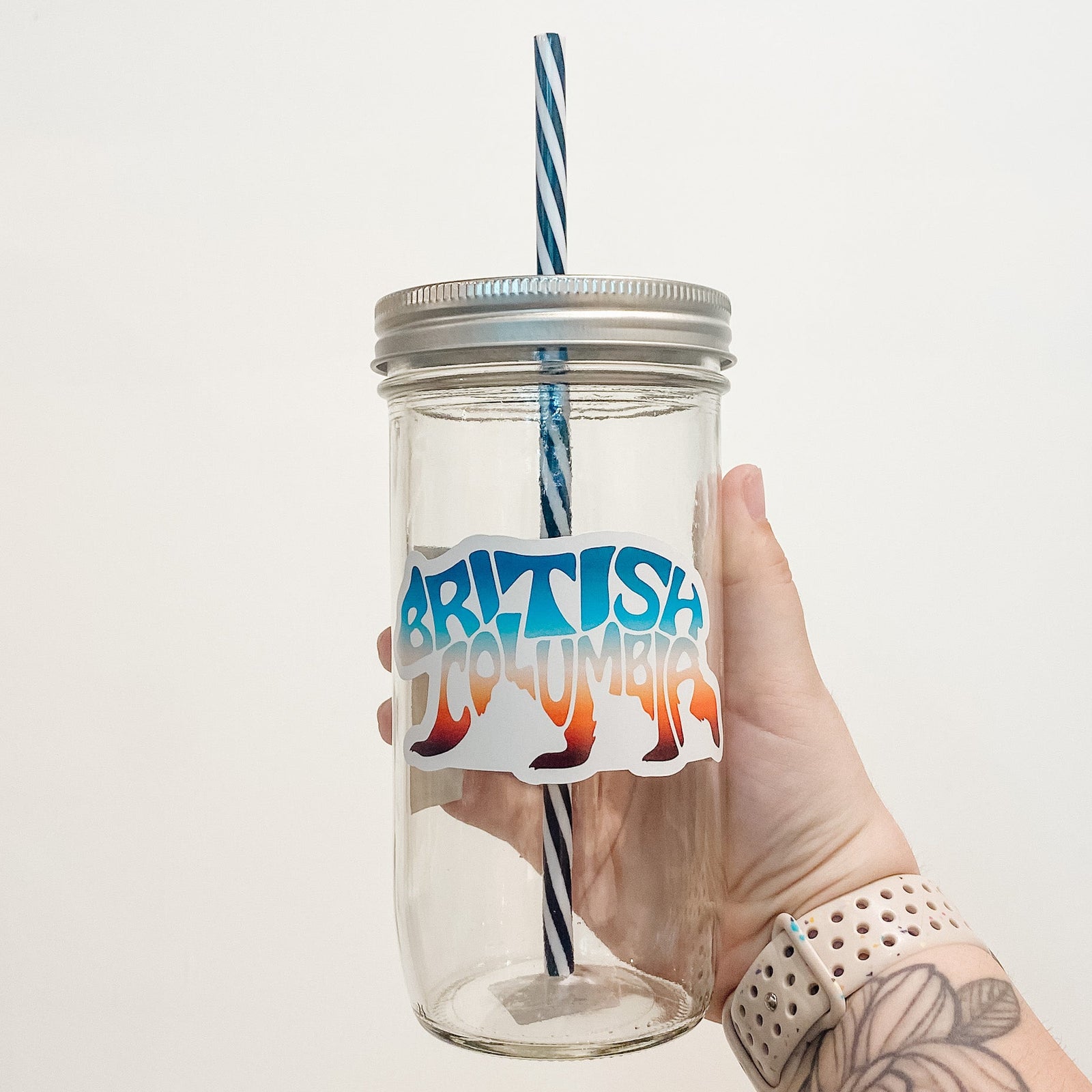 Hand holding mason jar that reads British Columbia in the shape of a bear. Colours are muted sunset gradient. 