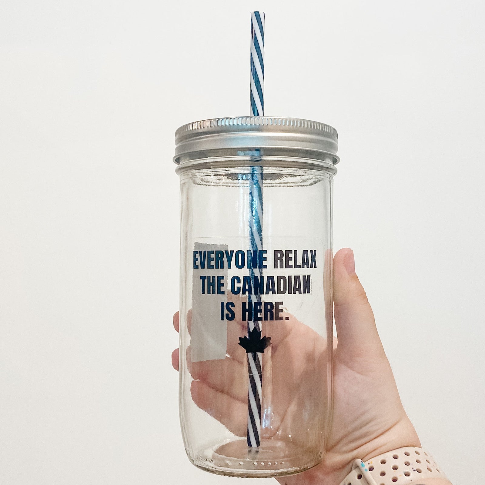 Hand holding mason jar that reads everyone relax the Canadian is here in black text. Small maple leaf at bottom. 