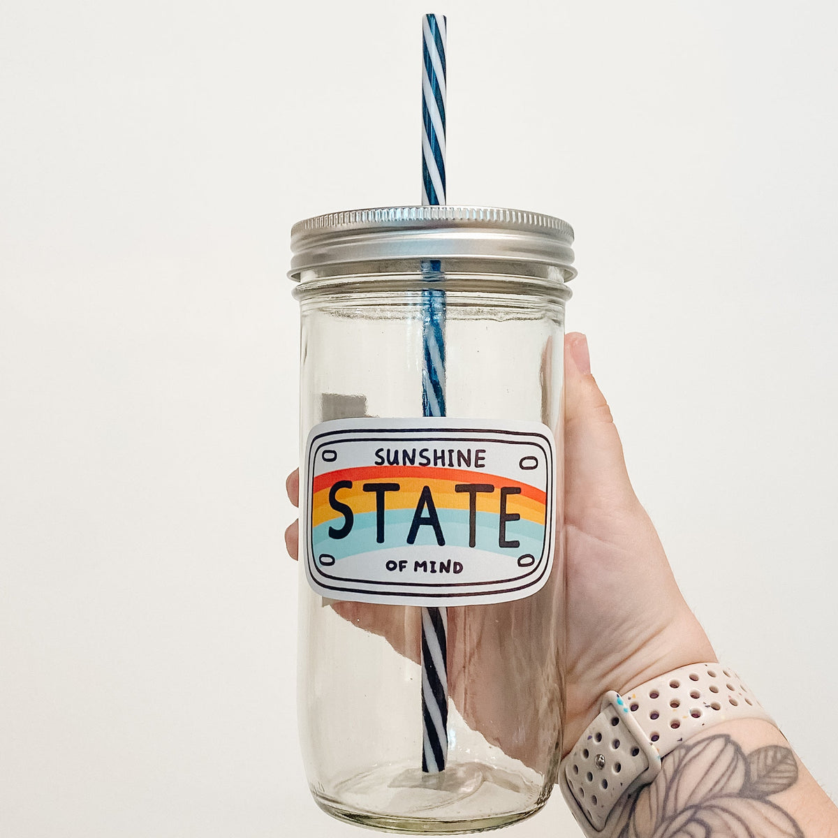 Hand holding mason jar that reads sunshine state of mind in the shape of a license plate with a rainbow across the middle.
