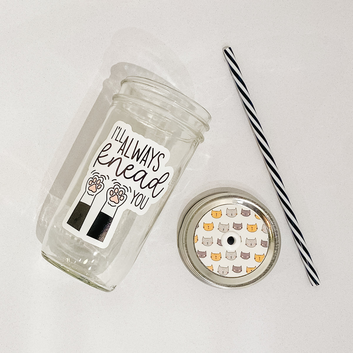 Flat lay of a mason jar tumbler that says “i&#39;ll always knead you”, a straw lid with different coloured cat faces, and a black and white striped straw.