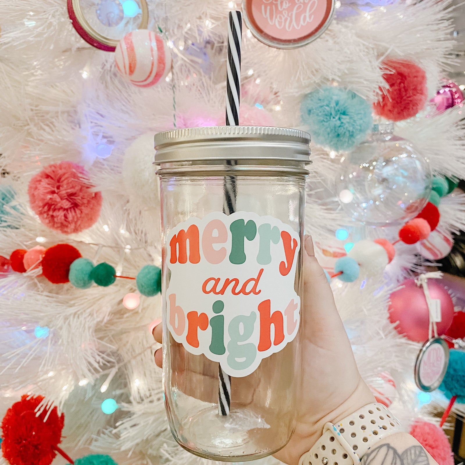 Hand holding mason jar Tumbler that reads Merry and Bright with blues, reds, pinks and teals for the font colors. Black and white striped plastic straw inside with Christmas tree in background.