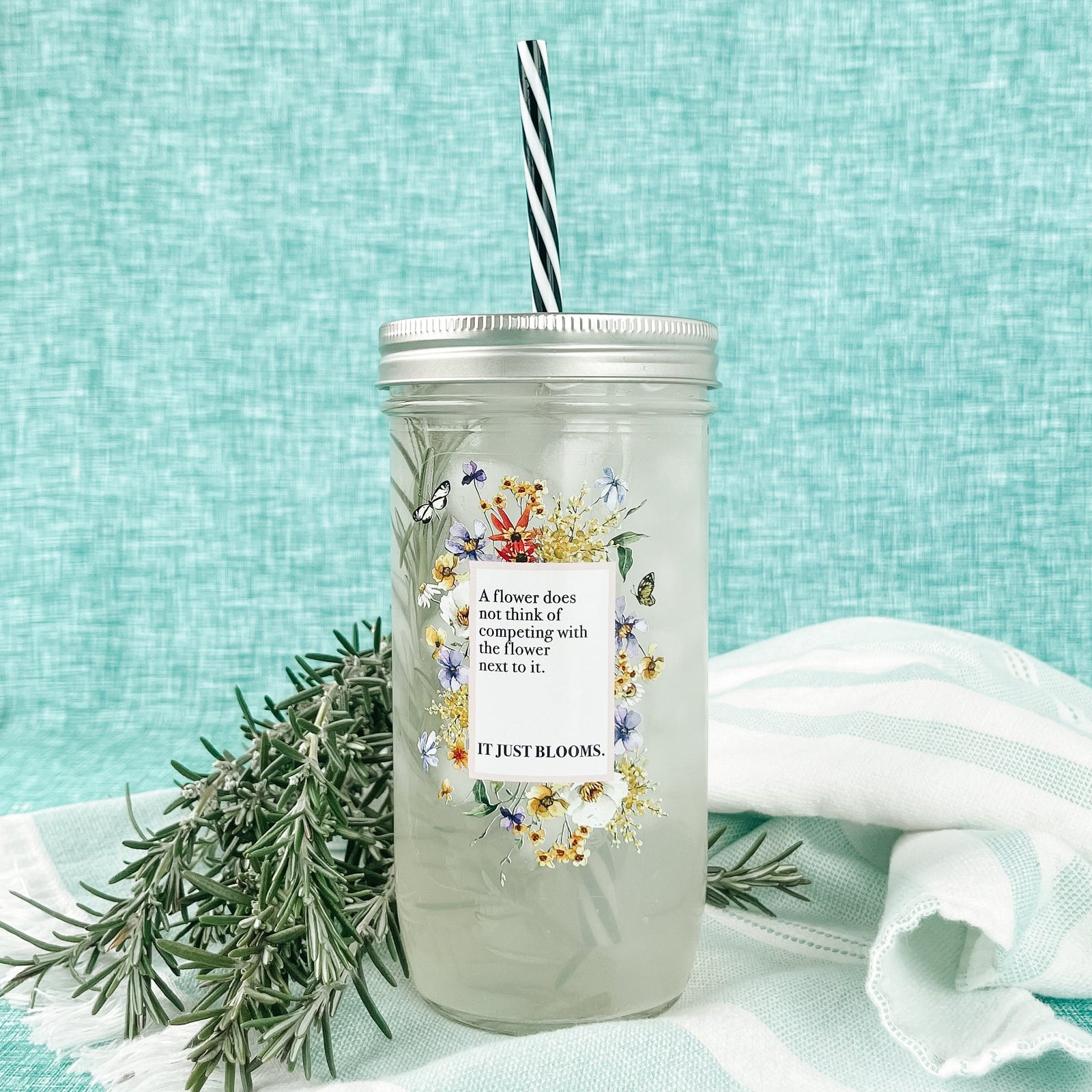 Mason jar tumbler that says “A flower does not think of competing with the flower next to it. It just blooms.” With flowers creating a rectangle around the words.