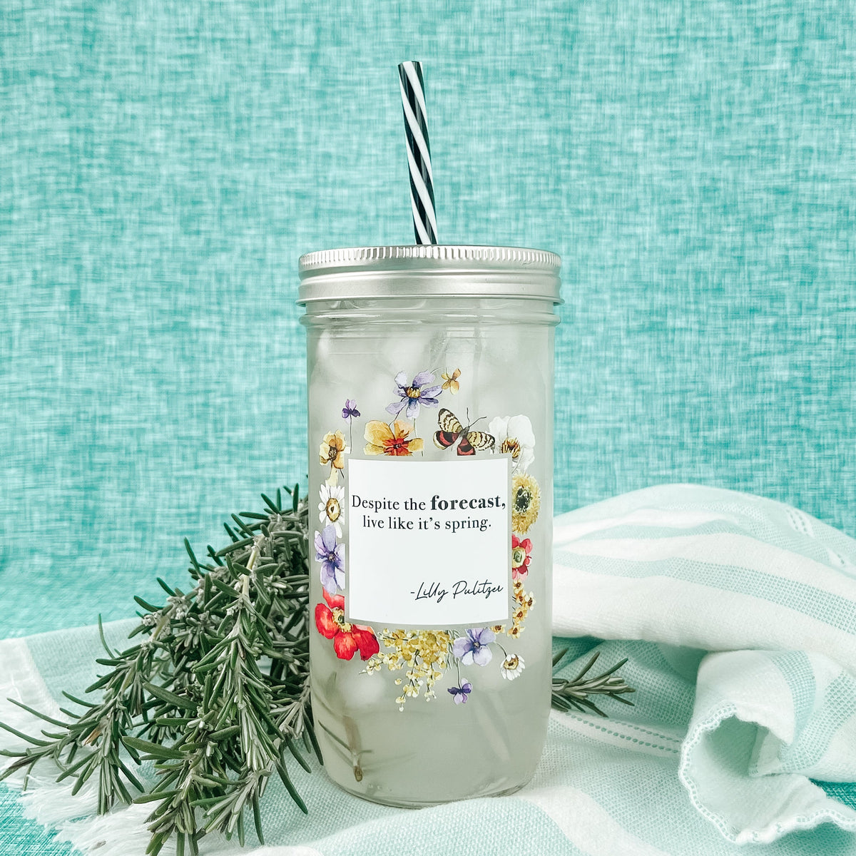 Mason jar tumbler that says “Despite the forecast, live like it’s spring. -Lilly Pulitzer” with flowers and butterflies creating a box around the words. 