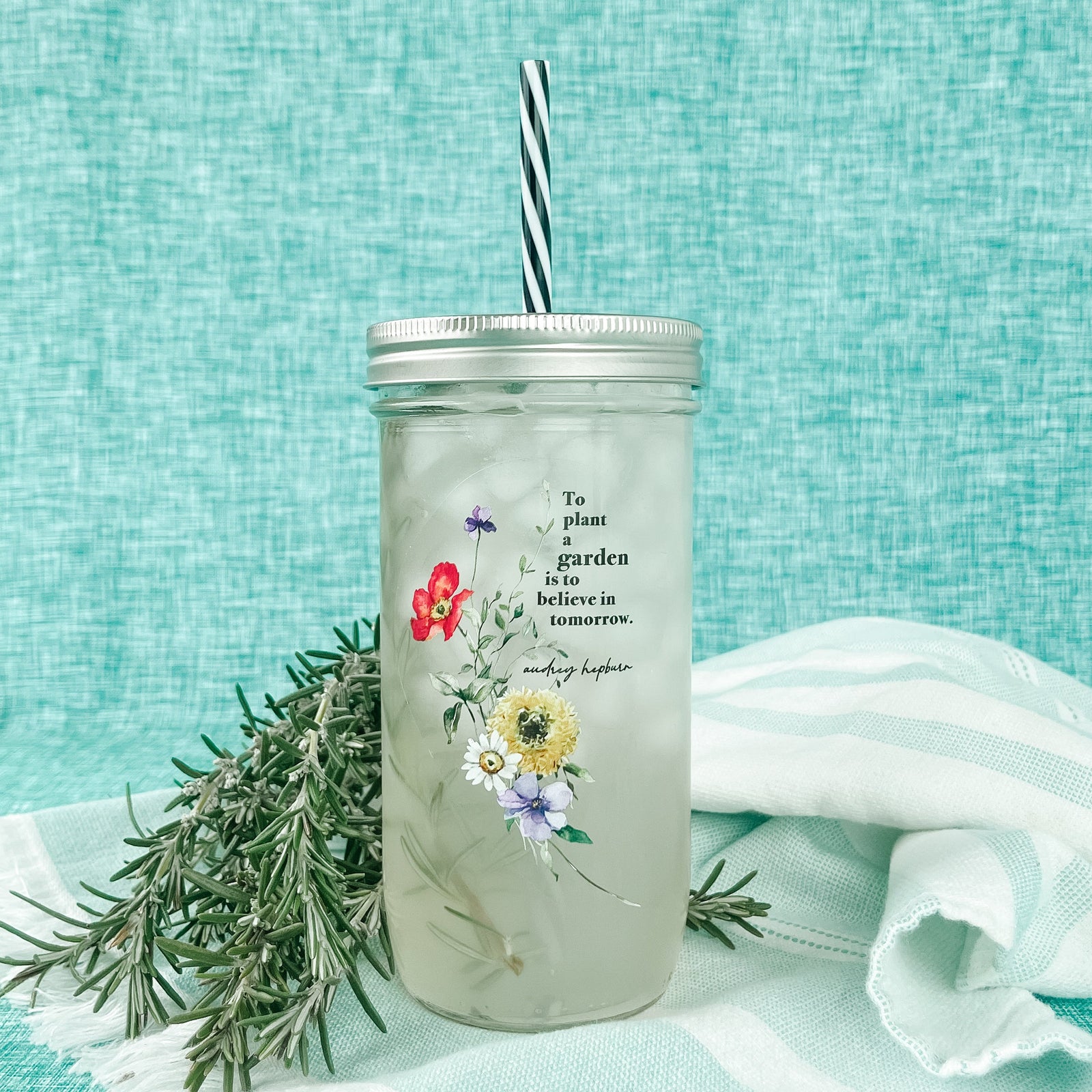 Mason jar tumbler that says “To plant a garden is to believe in tomorrow. Audrey Hepburn” with florals to the bottom left of the quote.