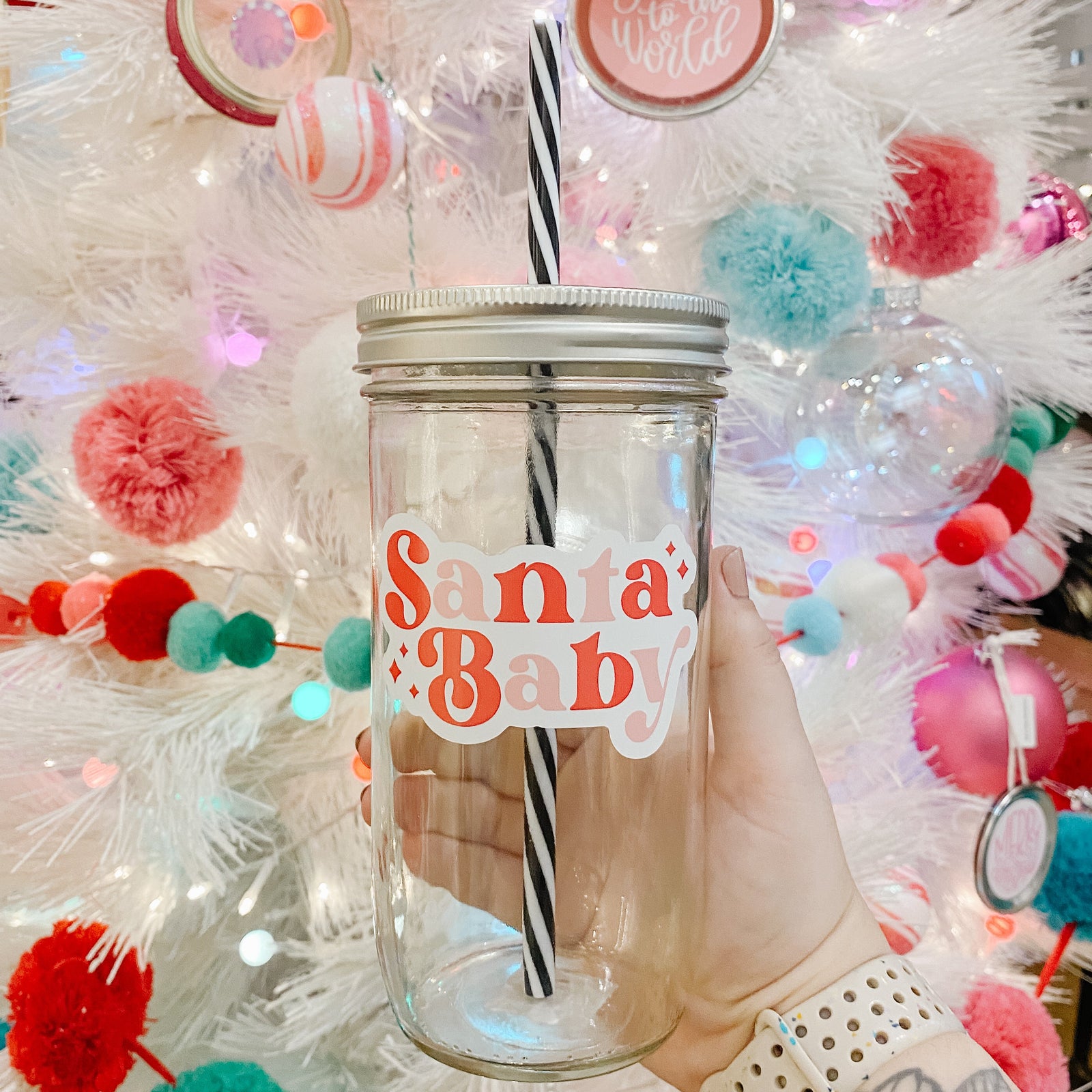 Hand holding mason jar Tumbler that reads Santa Baby with red and pinks for the font colors. Black and white striped plastic straw inside with Christmas tree in background.