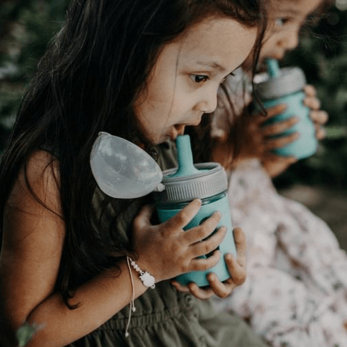 Two little girls drink out of a mason jar silicone squeeze pouch jar with a grey plastic ring and an aqua straw lid