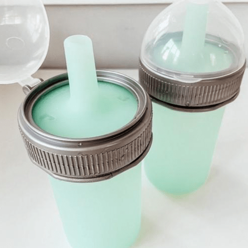 Two mason jar silicone squeeze pouch bottles with grey plastic rings and an aqua straw lid sit on a white background