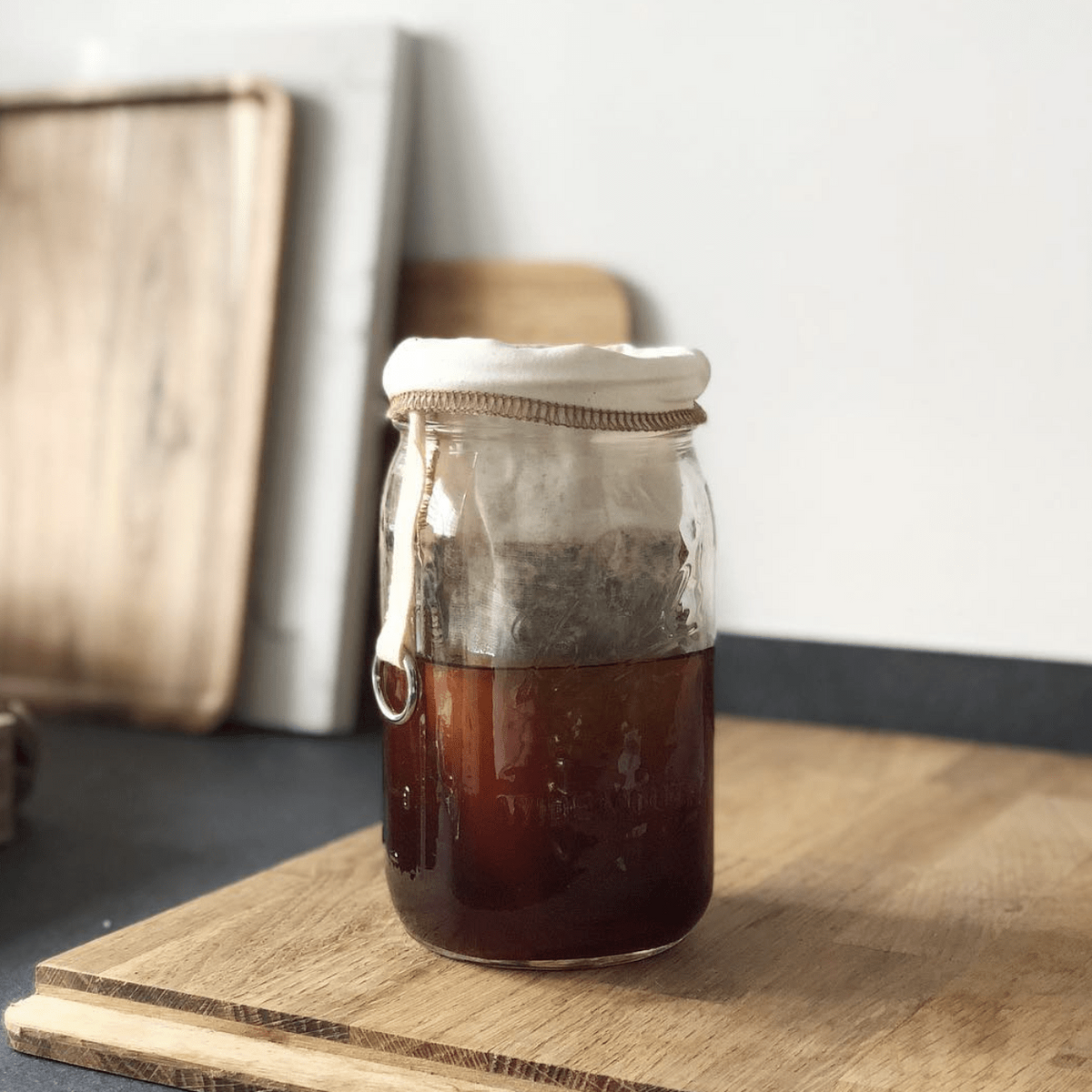 32 oz glass reusable mason jar sits on a wooden cutting board on a kitchen counter. The jar is filled with coffee and has a coffee filter with coffee ground in it