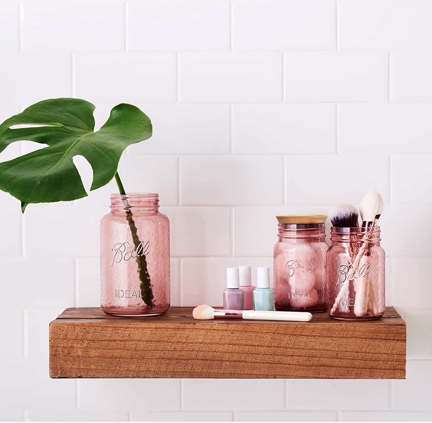 picture of a bathroom shelf with makeup, nail polish, and three mason jars that are a rose pink colour. Each jar has something inside it: a leaf, cotton balls, and makeup brushes