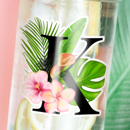 Transparent Tumbler with water and lemon inside and a sticker of flower, coconut leaf and ornamental plants with big letter K printed on it. Tumbler is photographed laying on a table with banana leaf and sunglasses beside it.