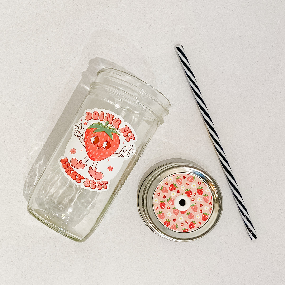 Flat lay of a mason jar tumbler that says “doing my berry best”, a light pink straw lid with strawberries (various shades of pink) covering it, and  black and white striped straw.