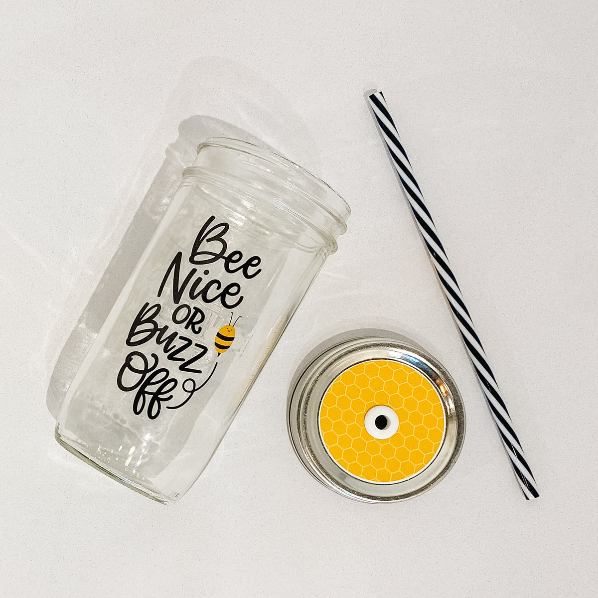Flat lay of a mason jar tumbler that says “bee nice or buzz off”, a straw lid that is yellow with light yellow hexagons to seem like a honeycomb, and a black and white striped straw. 
